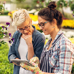 Adorable charming professional modern florist woman showing a list of flowers on a tablet to the curious attractive blonde female customer in a greenhouse.
