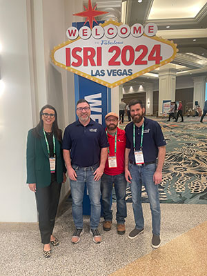 a woman and three men stand in front of an ISRI2024 sign