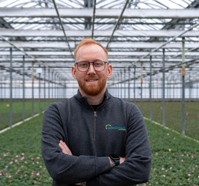 A headshot of Andrew Schooley, director of Venlo sales at Westbrook Greenhouse Systems, standing smiling with arms crossed inside a greenhouse filled with flowers.