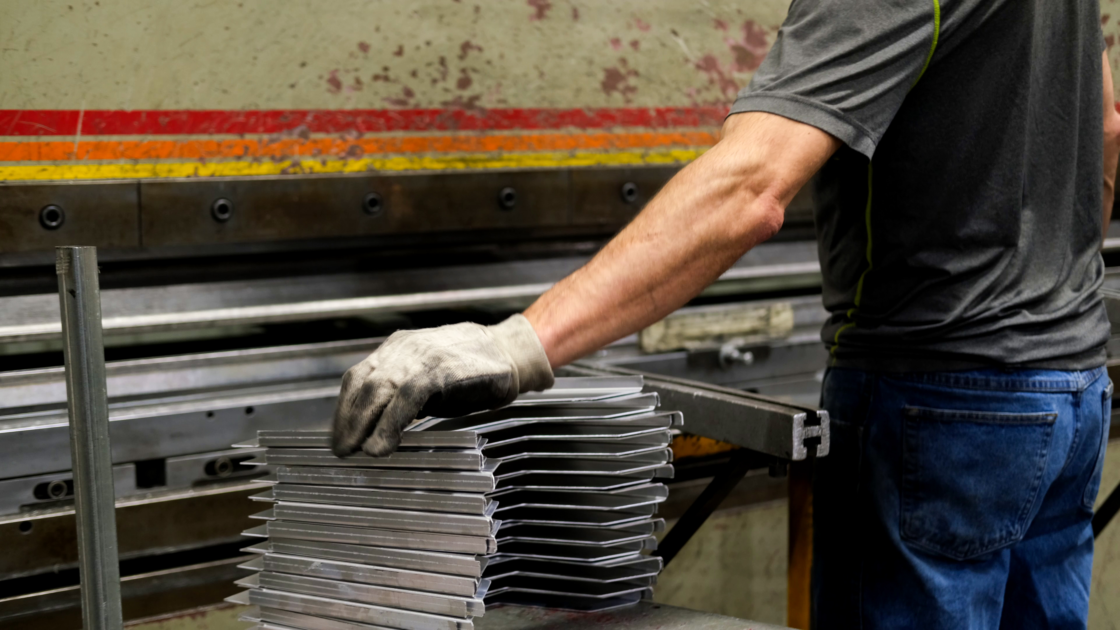A worker in a gray shirt and gloves stacks uniform metal components in front of a large industrial metal press inside Westbrook Greenhouse Systems’ manufacturing facility.