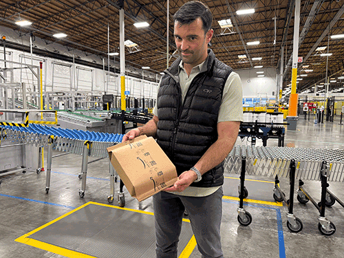 a white man with dark hair in a puffer vest holds an Amazon box